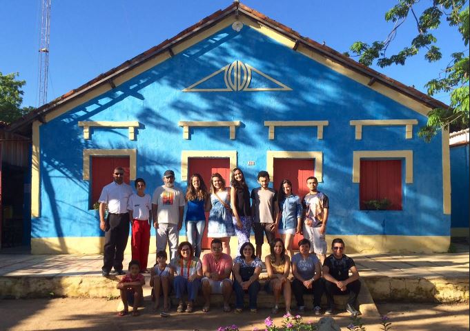  Foto da casa grande na cor azul celeste vista de frente, com a luz do sol refletida na mesma,  com quatro janelas vermelhas com moldura ao redor em gesso branco. Na frente da casa, na calada esto 17 pessoas, 8 homens e 9 mulheres. As pessoas posam na foto em duas filas, a primeira com 9 pessoas em p lado a lado e outra de 8 pessoas na frente sentadas lado a lado.